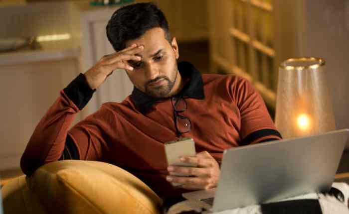 एक व्यक्ति सोफे पर बैठा मोबाइल देख रहा है, माथे पर हाथ रखकर सिर दर्द और थकान महसूस कर रहा है, पास में लैपटॉप खुला है. ज्यादा स्क्रीन टाइम और माइग्रेन का संकेत.